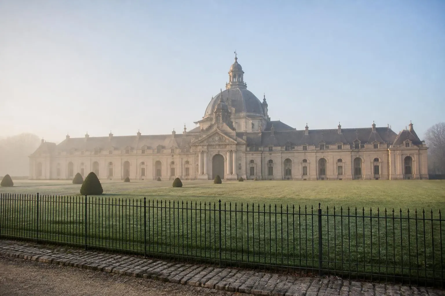 Façade et dôme d'un bâtiment d'écuries baroque en pierre claire au petit matin, pelouse et grille en fer forgé au premier plan