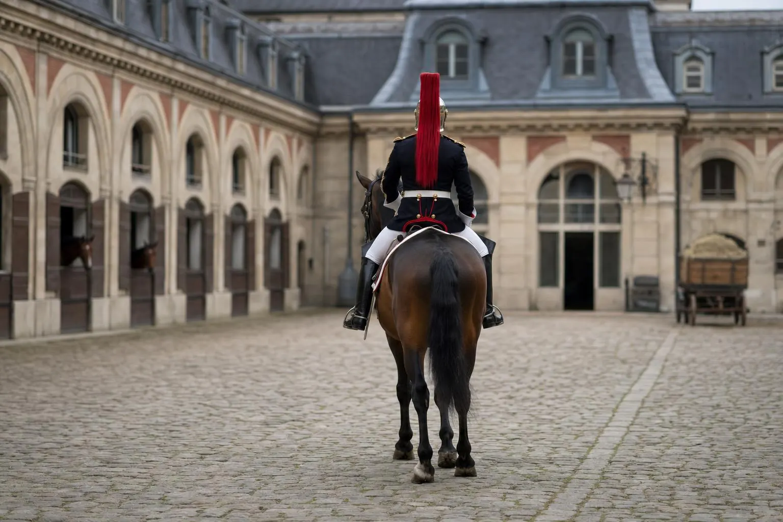 Cavalier de dos en tunique bleu nuit et casque à crinière rouge sur un cheval bai, cour pavée d'une caserne à arcades