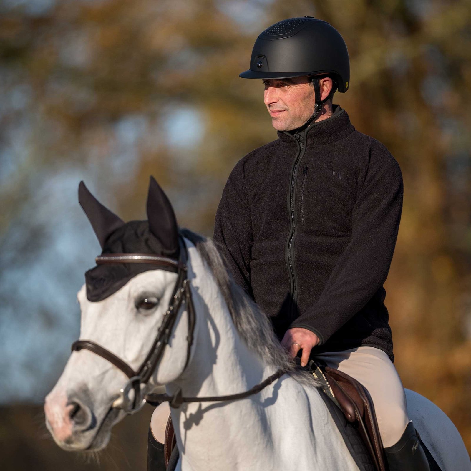 Cavalier portant le casque Strixx au pas sur un cheval gris avec bonnet noir, arbres d'automne en fond