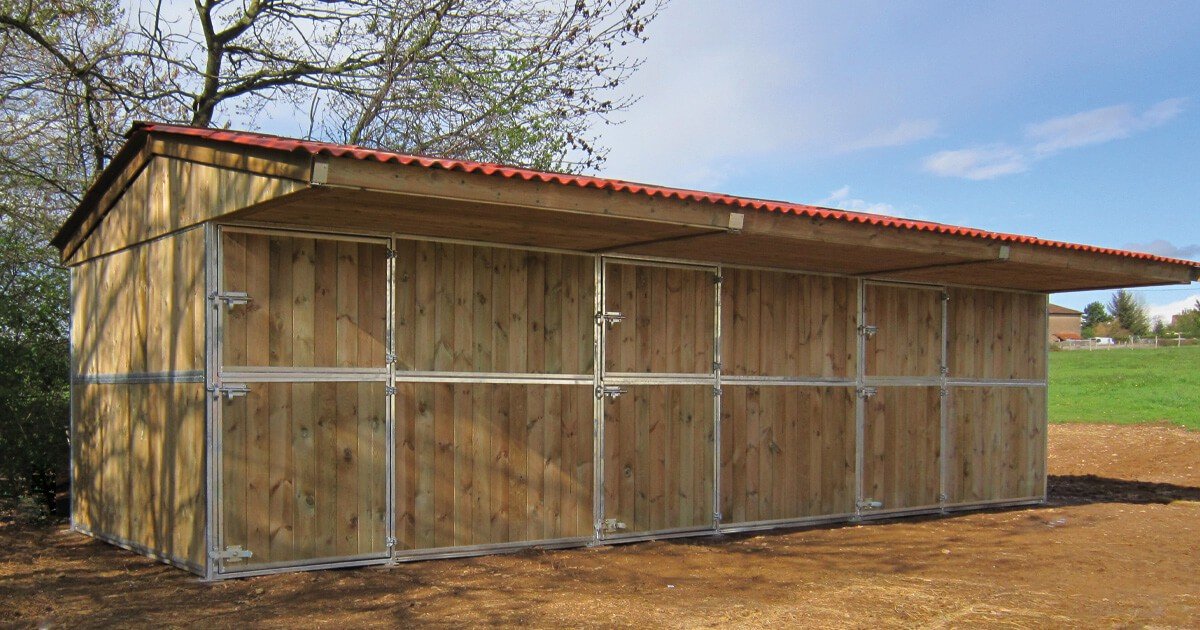 Abri-box Cheval Liberte en bois a toit rouge avec portes metalliques, installe sur un paddock