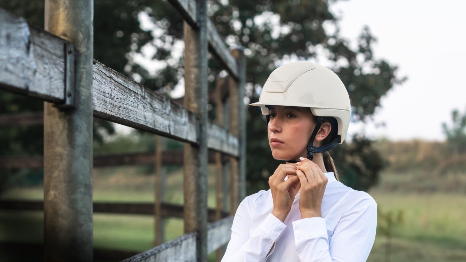 Cavalière en chemise blanche attachant la jugulaire d'un casque KASK Nemesi crème, appuyée à une barrière en