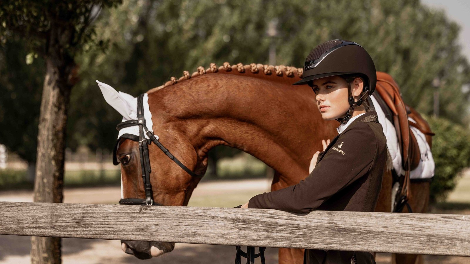 Cavalière portant un casque KASK marron, accoudée à une lice à côté de son cheval alezan tressé