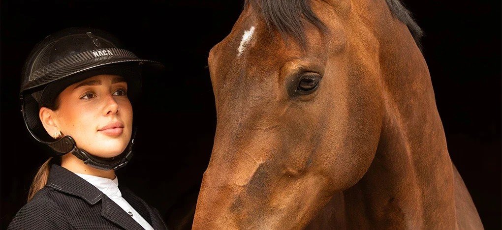 Cavalière portant un casque NACA noir à visière, joue contre la tête d'un cheval bai, fond noir