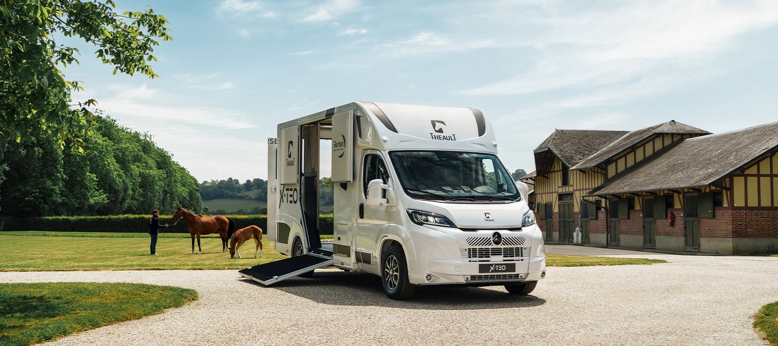 Van Theault XTEO blanc rampe baissée devant des écuries, chevaux au paddock